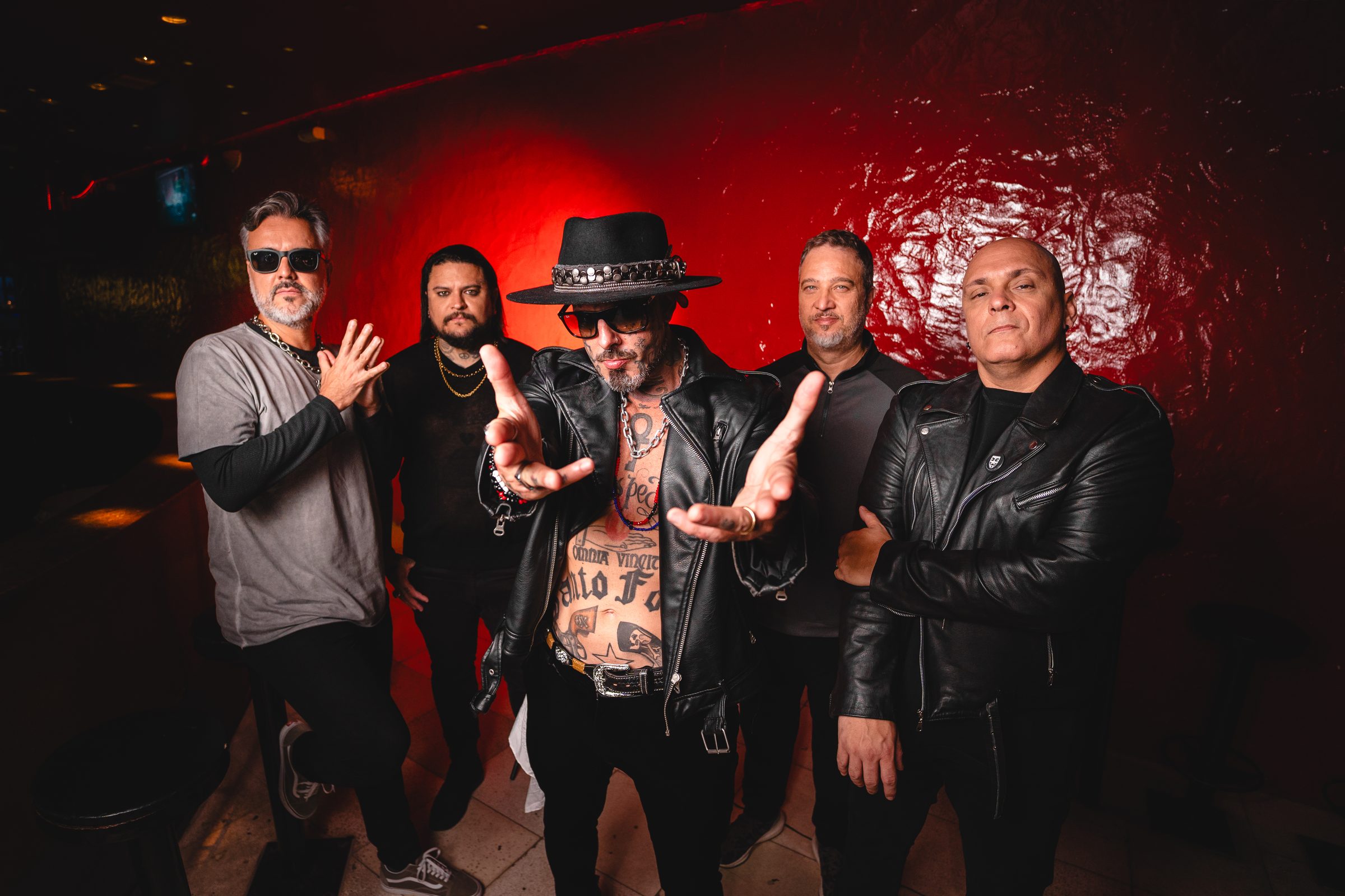 Os cinco integrantes do Detonautas diante de uma parede vermelha, o vocalista de chapéu com as mãos estendidas